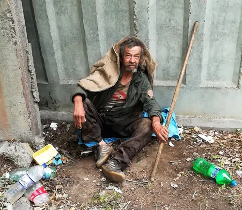 Бомжа Фото человек, сидящий на корточках, держит палку и бутылку воды