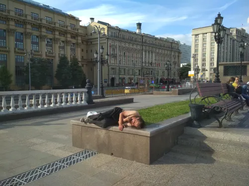 Бомжей Фото человек, лежащий на каменной скамейке в парке с постройками на заднем плане